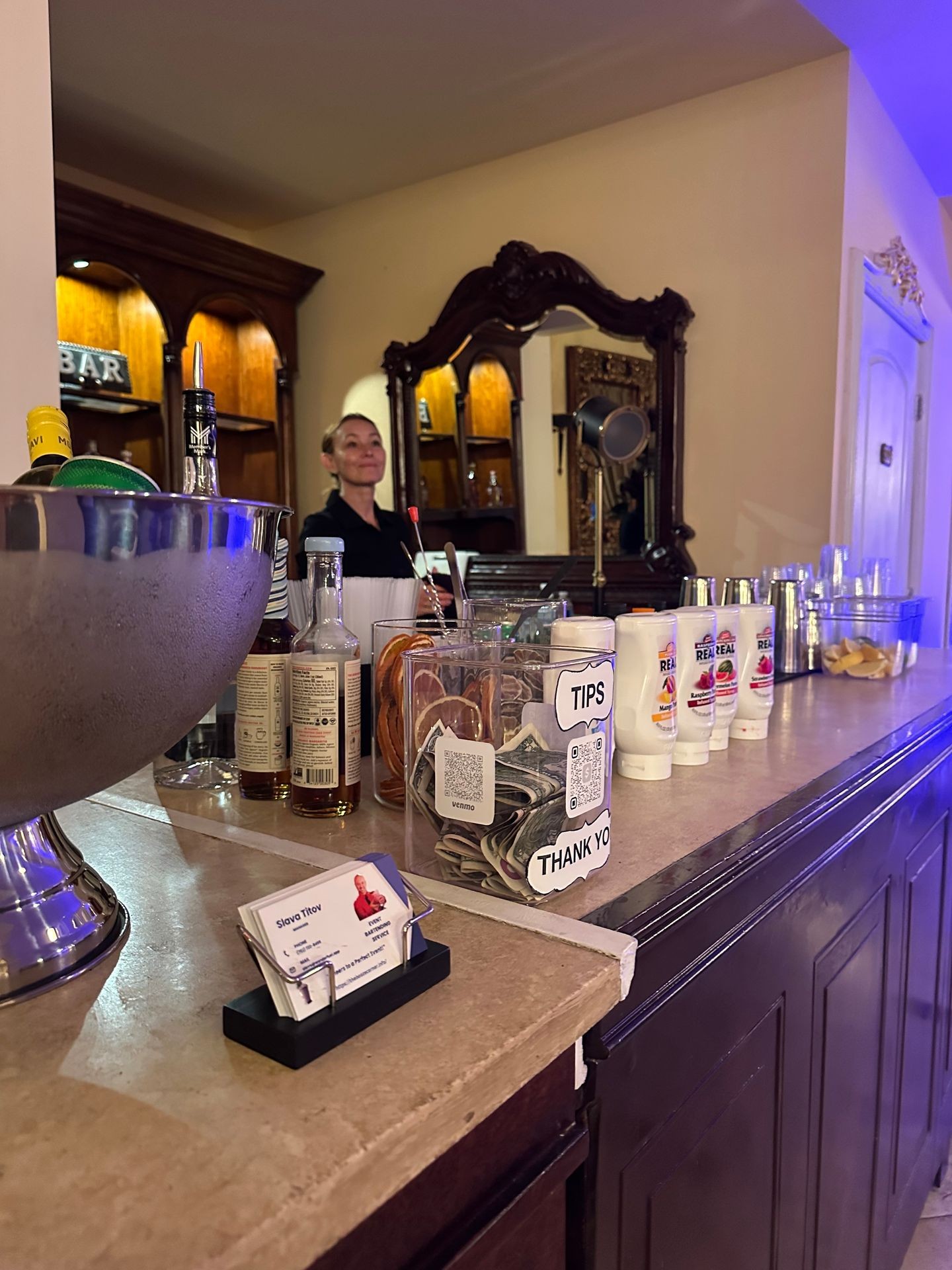 Bar counter with tip jar, bottles, napkins, and bartender in the background smiling.