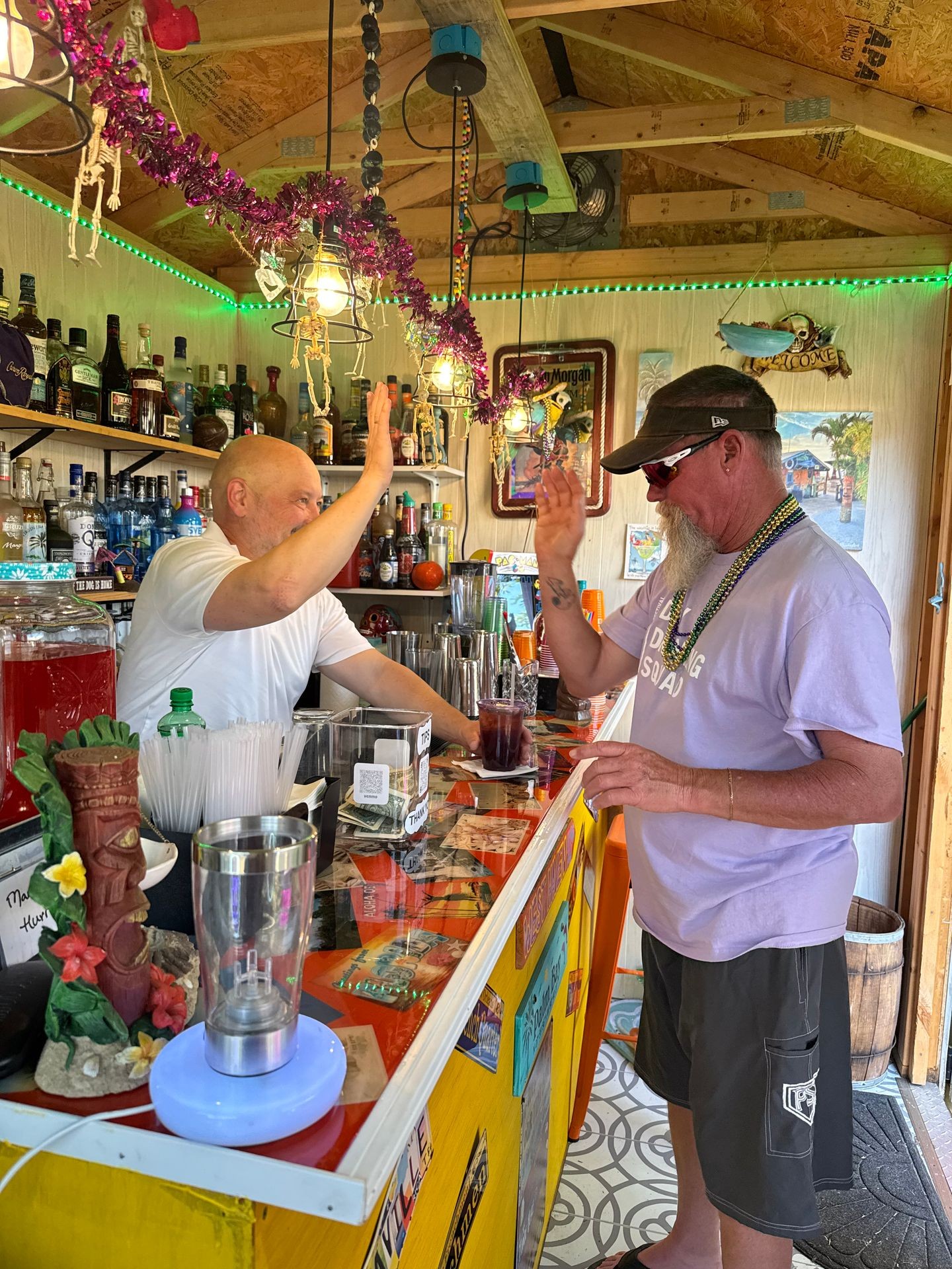 Two people high-fiving at a colorful bar with decorated shelves and hanging lights.